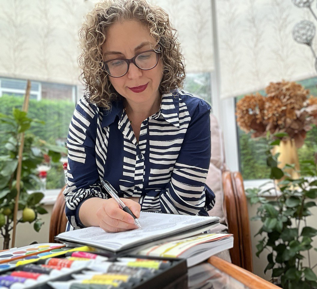 Sanaz is drawing a pattern, doodling floral designs to transfer to Adobe Illustrator for the final pattern and colouring it. Sitting in the conservatory with all plants and flowers around her. And a box of watercolour tubes on the table.